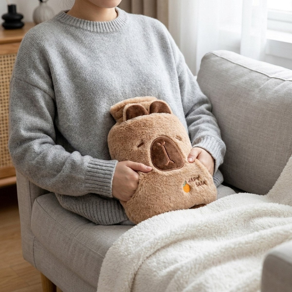 Bouillotte peluche capybara sur les genoux, mains glissées dans ses poches, sur un canapé gris