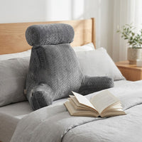 Coussin de lecture à accoudoirs gris contre la tête d’un lit, livre ouvert sur la couette — mise en situation illustrative